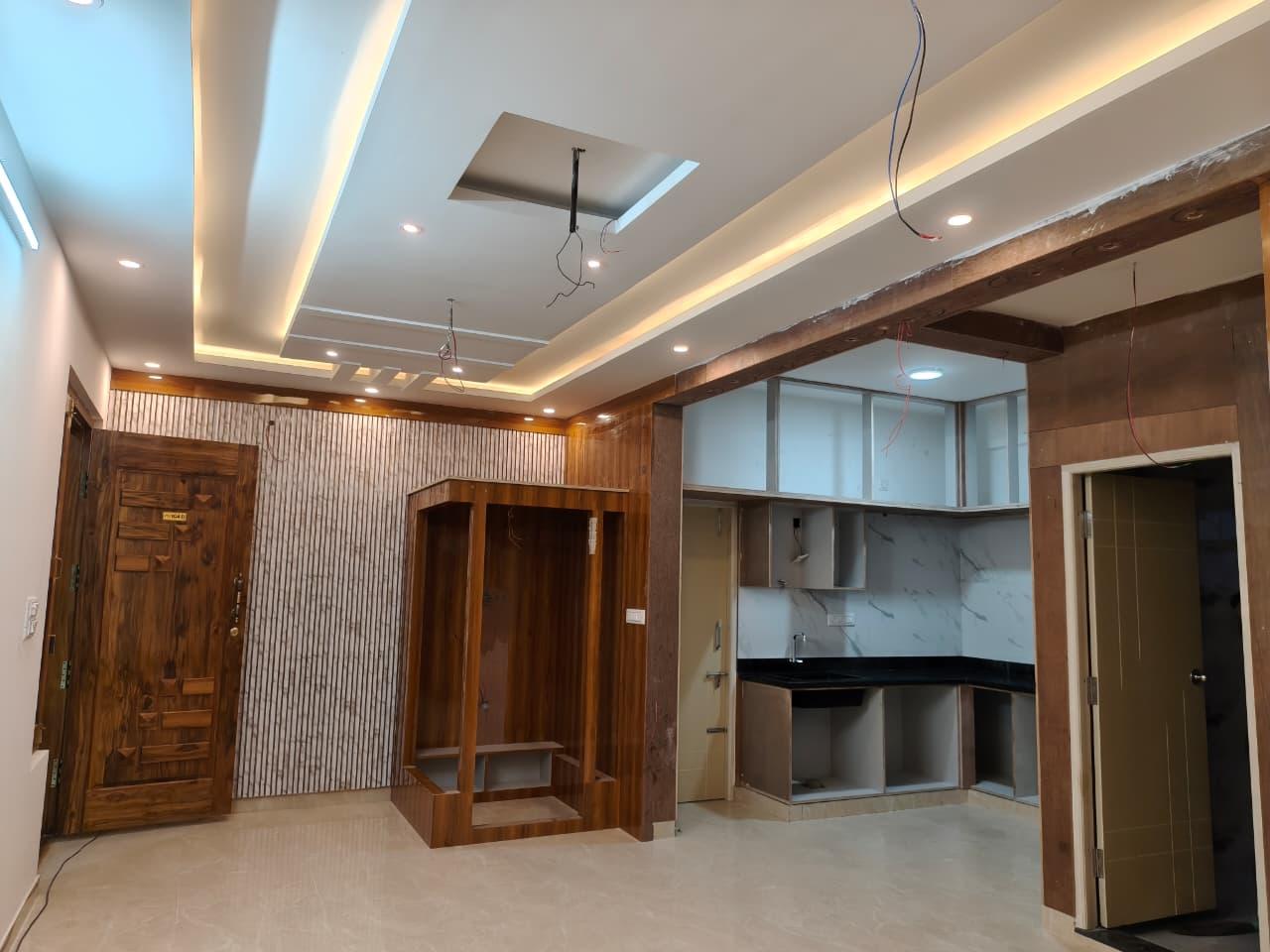 A wide view of a flat during fit-out, showing a false ceiling with cove lighting, timber wall panelling, a built-in storage loft and the kitchen, with trades still to complete.