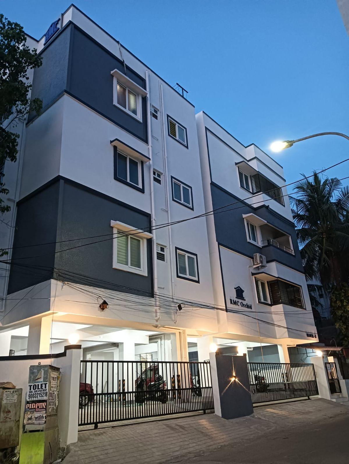A modern apartment community block finished in white and grey, seen at dusk with palm trees along the approach road
